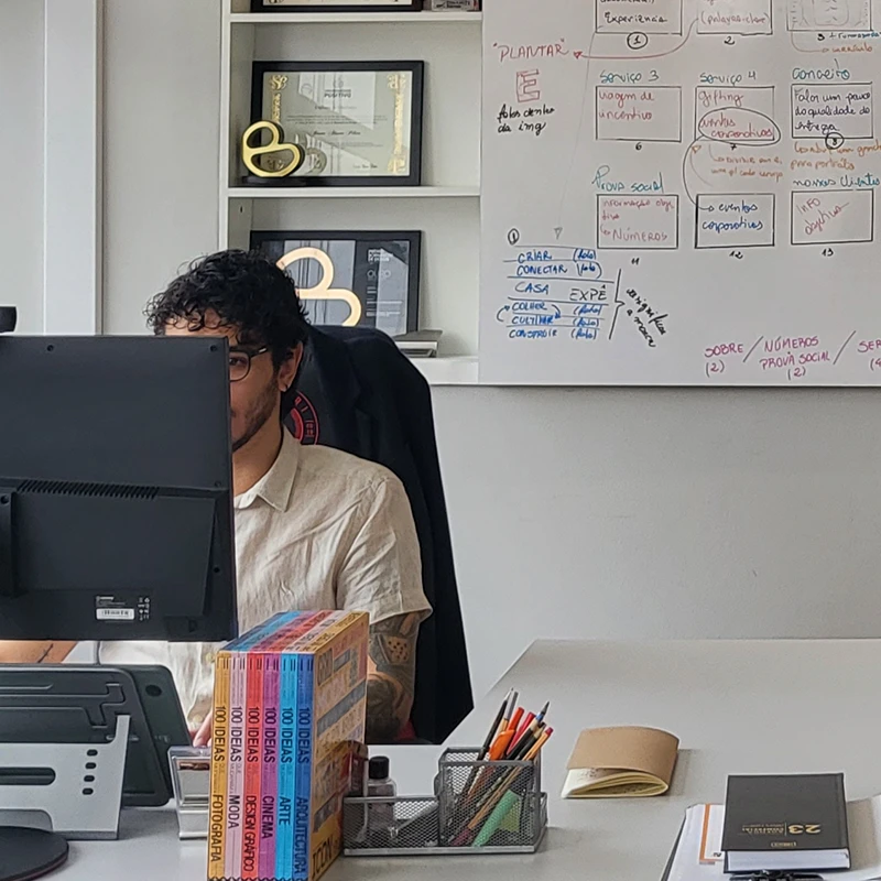 Homem com camisa clara concentrado em computador em escritório moderno, cercado por prêmios e cadernos em mesa organizada.